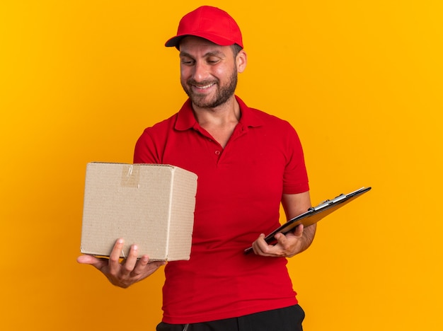 Souriant jeune livreur caucasien en uniforme rouge et casquette tenant une boîte en carton et un presse-papiers regardant une boîte en carton isolée sur un mur orange