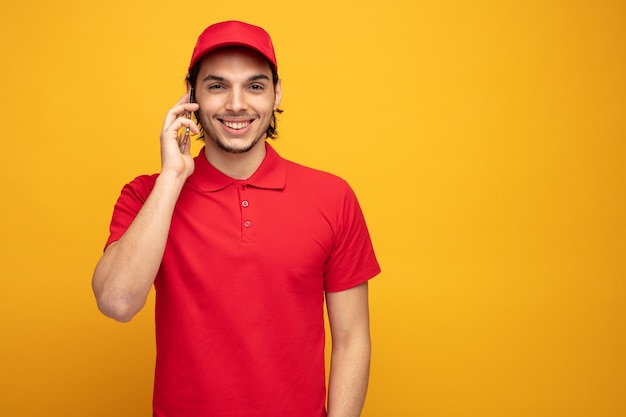 souriant jeune livreur portant l'uniforme et la casquette regardant la caméra tout en parlant au téléphone isolé sur fond jaune avec espace de copie