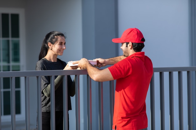 Souriant jeune livreur en uniforme rouge tenant une boîte donne à la belle femme costumière devant la maison.