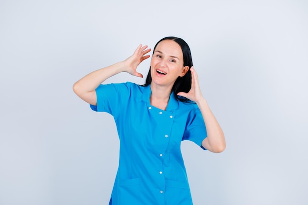 Souriant jeune médecin regarde la caméra en se tenant la main près des cheveux sur fond blanc