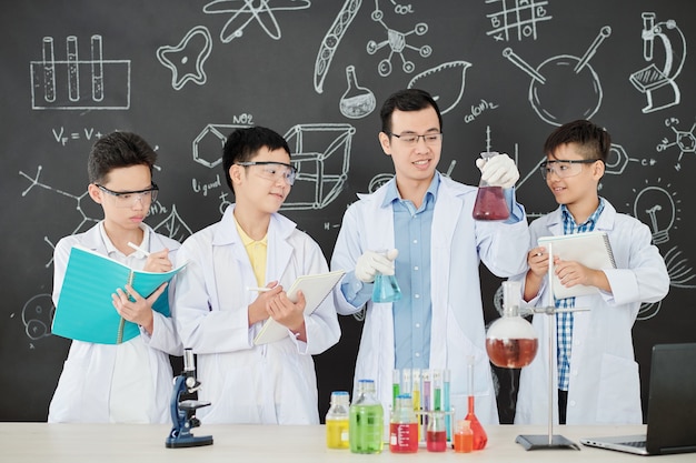 Souriant jeune professeur de chimie asiatique montrant quels liquides il va mélanger pour montrer une réaction chimique aux étudiants