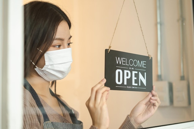 Souriant jeune propriétaire asiatique détaillant, femme de café tournant le panneau d'affichage pour ouvrir le masque facial, protéger contre la pandémie de coronavirus, rouvrir le magasin après avoir fermé la quarantaine de verrouillage dans covid à la nouvelle normale.