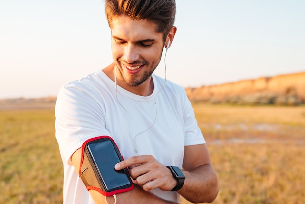 Souriant jeune sportif à l'aide de téléphone mobile à écran blanc sur le brassard à l'extérieur