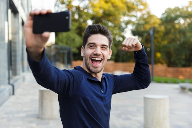 Souriant jeune sportif prenant un selfie assis à l'extérieur, posant