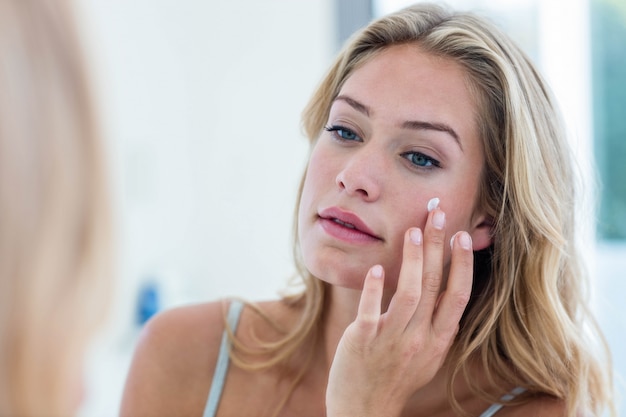 Souriant jolie femme appliquant la crème sur son visage dans la salle de bain