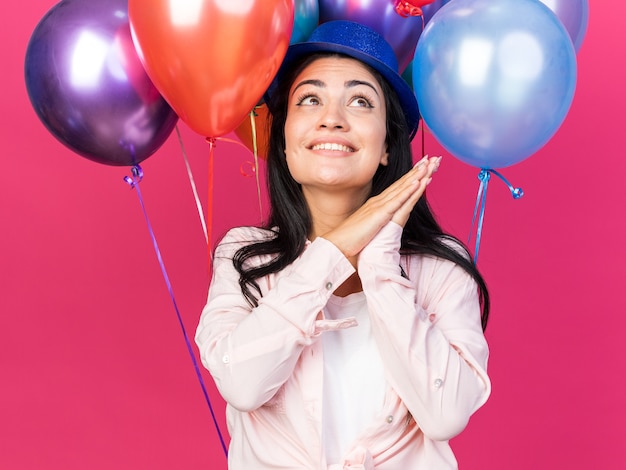 Souriant en levant la belle jeune fille portant un chapeau de fête debout devant des ballons tenant les mains ensemble isolés sur un mur rose