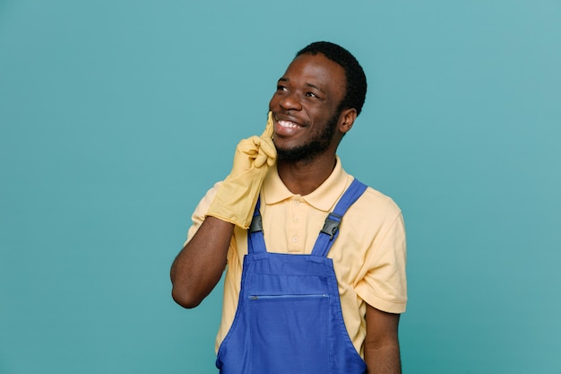 Souriant Mettant Le Doigt Sur La Joue Jeune Homme Nettoyeur Afro-américain En Uniforme Avec Des Gants Isolés Sur Fond Bleu