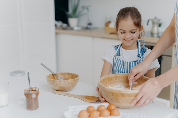 Souriant petit assistant fille tient grand bol, regarde comment mère mélange des œufs avec de la farine