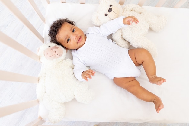 Souriant petit bébé afro-américain dans un lit blanc avec des jouets ours