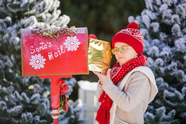 Souriant petit garçon avec un chapeau rouge et des lunettes vertes avec sa lettre près de la boîte aux lettres du père Noël