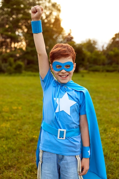 Photo souriant préadolescent intrépide portant un masque de super-héros bleu et une cape debout avec le bras levé et célébrant le succès tout en regardant la caméra