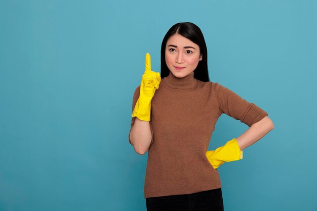 Souriant satisfait ravi femme de ménage asiatique portant des gants jaunes et pointant le doigt vers le haut pour copier l'espace, concept de maison de nettoyage, femme de ménage optimiste et positive de Glade