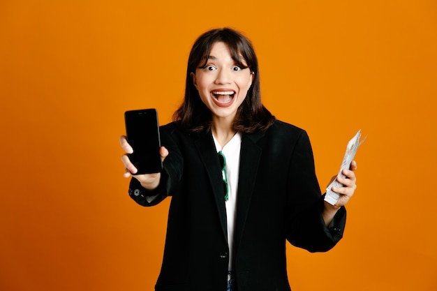 souriant tenant de l'argent et téléphone jeune belle femme portant une veste noire isolée sur fond orange