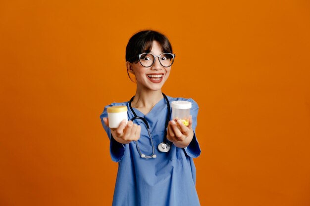 Souriant tenant des contenants de pilules à la caméra jeune femme médecin portant un stéthoscope fith uniforme isolé sur fond orange