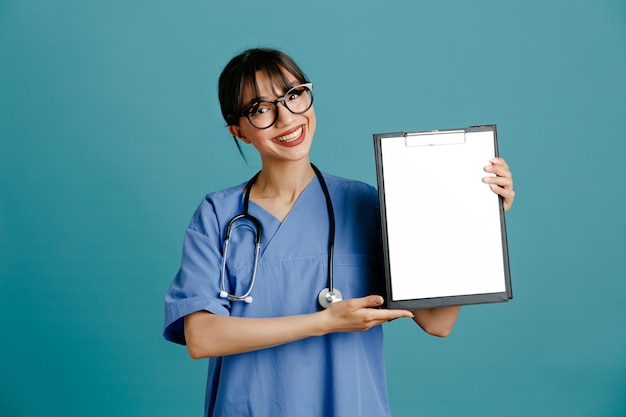 Souriant tenant le presse-papiers jeune femme médecin portant un stéthoscope fith uniforme isolé sur fond bleu