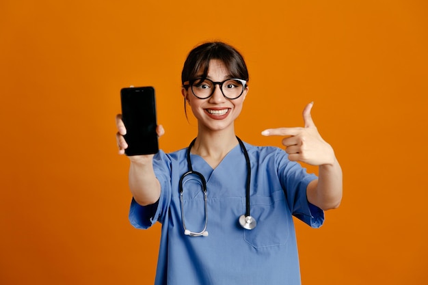 Souriant tenant le téléphone à la caméra jeune femme médecin portant un stéthoscope fith uniforme isolé sur fond orange