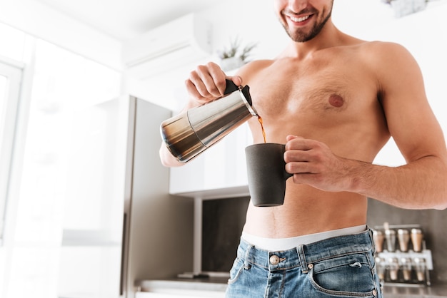 Souriant torse nu jeune homme debout et verser du café dans la tasse sur la cuisine