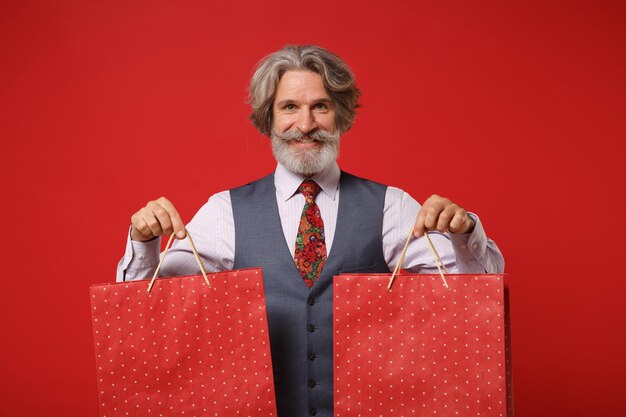 Souriant vieil homme barbu moustache aux cheveux gris en chemise gilet cravate posant isolé sur fond rouge. Concept de style de vie des gens. Maquette de l'espace de copie. Tenir le sac d'emballage avec les achats après le shopping.