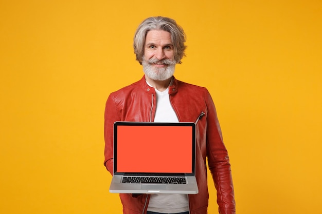 Souriant vieil homme barbu moustache aux cheveux gris en veste de cuir isolé sur fond orange jaune. Concept de style de vie des gens. Maquette de l'espace de copie. Tenez un ordinateur portable avec un écran vide vide.