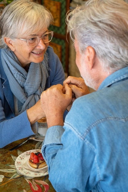 Souriant vieux couple de personnes âgées mains dans les mains regardant dans les yeux au café. Cappuccino et gâteau aux fruits sur la table