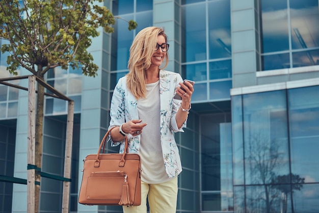 Souriante belle blogueuse à la mode dans des vêtements élégants et des lunettes avec un sac à main, à l'aide d'un smartphone, debout contre un gratte-ciel.