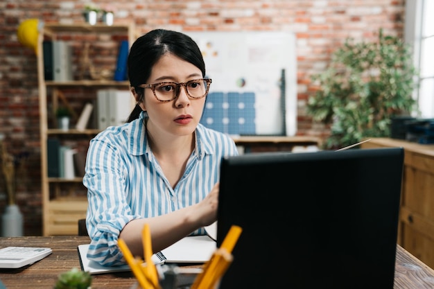 Souriante belle femme d'affaires chinoise asiatique assise à un bureau en bois dans un bureau moderne confortable regardant sérieusement l'écran d'ordinateur portable travaillant et la verdure dans le mur de briques rouges. respectueux de la nature