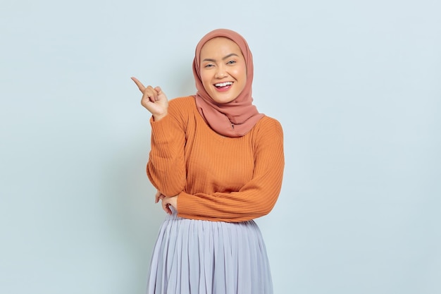 Souriante belle femme asiatique en pull marron et hijab pointant le doigt dans l'espace de copie montrant des produits publicitaires et confiant regardant la caméra isolée sur fond blanc