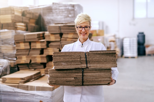 Souriante belle femme blonde relocalisant des boîtes de démontage en entrepôt.