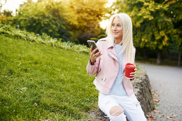 Souriante belle femme écoutant de la musique relaxante dans les écouteurs avec son téléphone portable ou sa tablette dans le parc. l'étudiant écoute une conférence en ligne et se prépare aux examens.