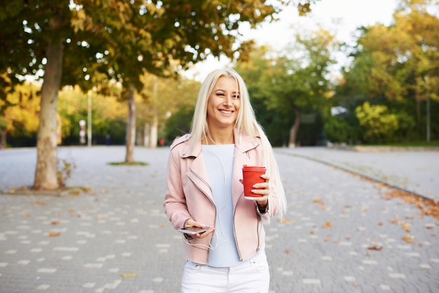 Souriante belle femme écoutant de la musique relaxante dans les écouteurs avec son téléphone portable ou sa tablette dans le parc. l'étudiant écoute une conférence en ligne et se prépare aux examens.