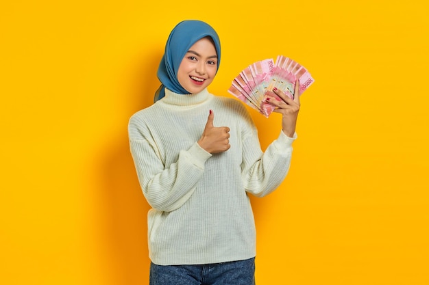 Souriante belle femme musulmane asiatique en pull blanc montrant de l'argent en espèces en billets de roupie indonésienne gesticulant pouces vers le haut isolé sur fond jaune Concept de style de vie religieux des gens