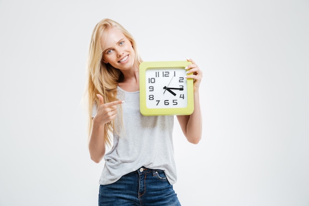 Souriante belle fille blonde pointant le doigt sur l'horloge murale isolée sur un mur blanc