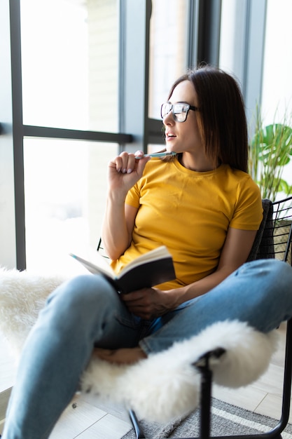 Souriante belle fille écrivant dans un journal tout en étant assis sur une chaise dans le salon