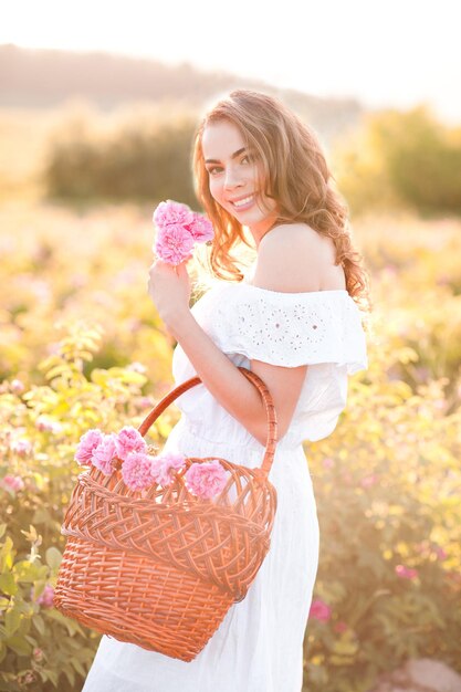 Souriante belle fille vêtue d'une robe blanche tenant un panier avec des roses à l'extérieur