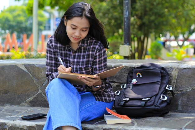 souriante belle jeune femme asiatique profiter d'écrire à faire liste idée dans le cahier dans le parc de la ville en plein air