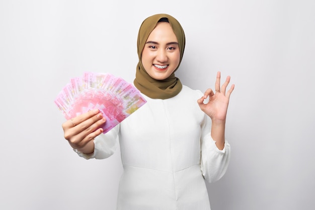 Souriante belle jeune femme musulmane asiatique portant le hijab détenant de l'argent en billets de roupie à la main et montrant le signe ok ok isolé sur fond blanc Concept de style de vie religieux des gens