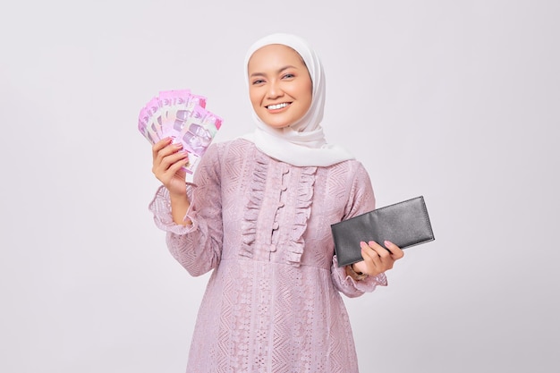 Souriante belle jeune femme musulmane asiatique portant le hijab et la robe violette tenant un portefeuille noir et des billets en roupies en espèces isolés sur fond de studio blanc