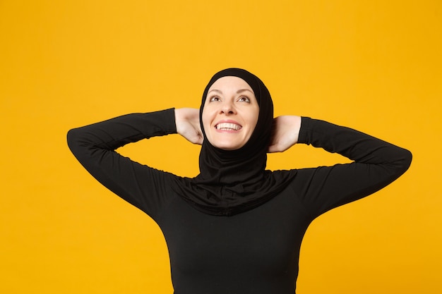 Souriante confiante belle jeune femme musulmane arabe en vêtements noirs hijab posant isolé sur mur jaune, portrait. Concept de mode de vie de l'Islam religieux.