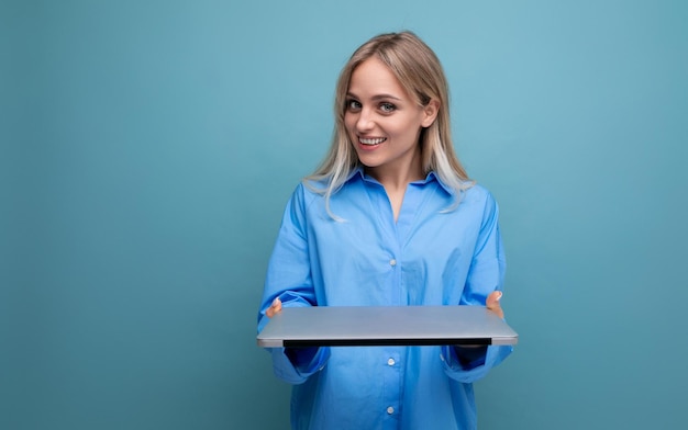 Souriante étudiante blonde joyeuse avec un ordinateur portable sur un fond bleu clair