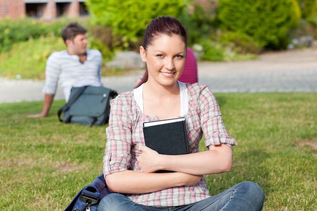 Souriante étudiante en lisant un livre assis sur l&#39;herbe