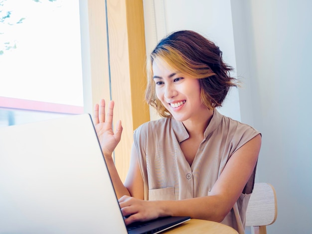 Souriante femme asiatique cheveux courts avec une mode tendance en surbrillance assise et utilisant un ordinateur portable ayant un appel vidéo Une femme adulte séduisante dit bonjour avec la main levée facetime réunion à la maison