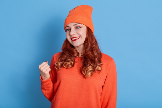 Souriante femme aux cheveux rouges serre les poings, porte du rouge à lèvres, porte un pull et un chapeau décontractés