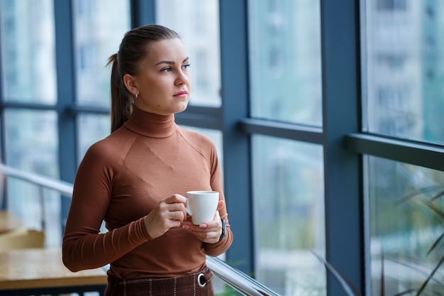 Souriante femme directrice heureuse pensant à son développement de carrière réussi tout en se tenant debout avec une tasse de café aromatique dans son bureau près de l'arrière-plan d'une fenêtre avec espace de copie