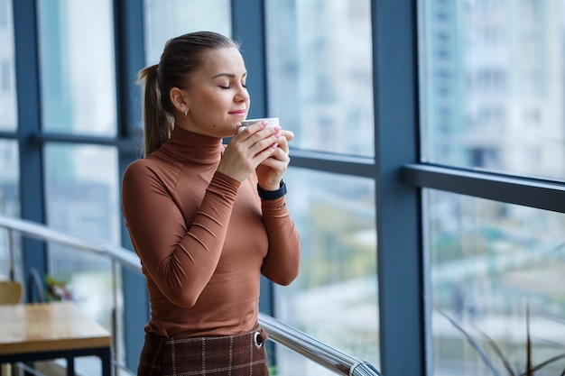 Souriante femme directrice heureuse pensant à son développement de carrière réussi tout en se tenant debout avec une tasse de café aromatique dans son bureau près de l'arrière-plan d'une fenêtre avec espace de copie