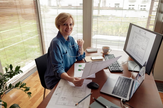 Souriante femme mature ingénieur constructeur travaille sur un projet depuis son bureau à domicile à l'aide d'un ordinateur