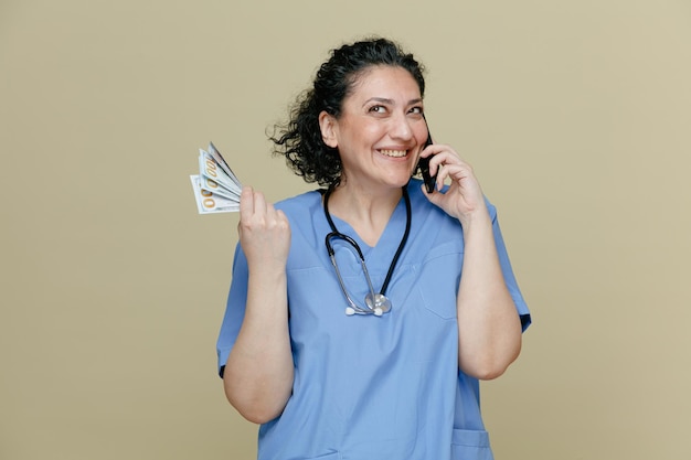 Souriante femme médecin d'âge moyen portant un uniforme et un stéthoscope autour du cou tenant de l'argent en levant les yeux tout en parlant au téléphone isolé sur fond olive