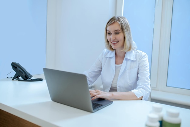 Souriante femme médecin concentrée dans la blouse blanche en tapant sur l'ordinateur portable de son bureau