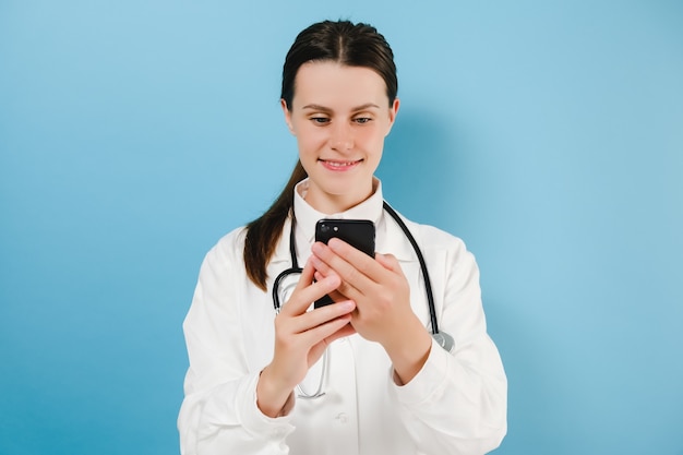 Souriante femme médecin mignonne en blouse blanche debout isolée sur fond bleu tenir le smartphone, vérifier les messages du patient, utiliser l'application mobile pour les travailleurs médicaux utilisation facile du confort du concept de technologie moderne