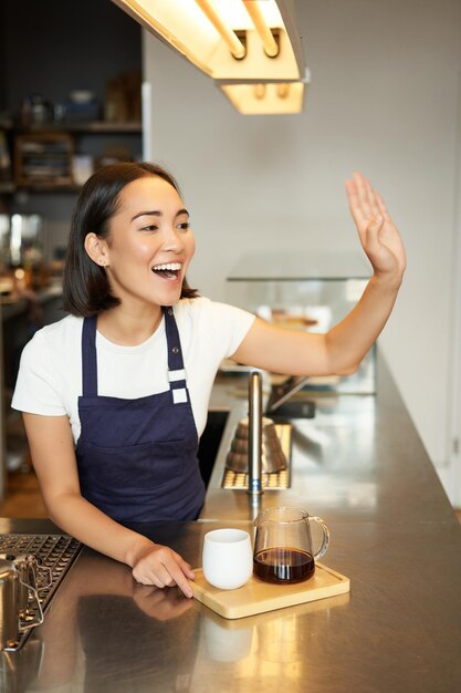 Souriante fille asiatique barista appelant le client donnant la commande dans un café préparé par lots infuser du café filtre