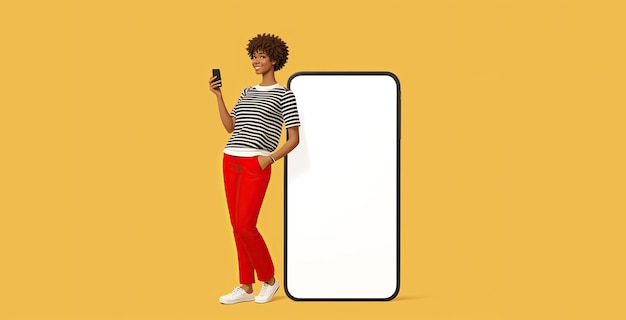 Photo souriante heureuse jeune femme afro-américaine, elle se tient près d'une maquette de téléphone portable mobile à grand écran vierge avec une zone de maquette d'espace de copie utilise un smartphone isolé sur un fond jaune uni